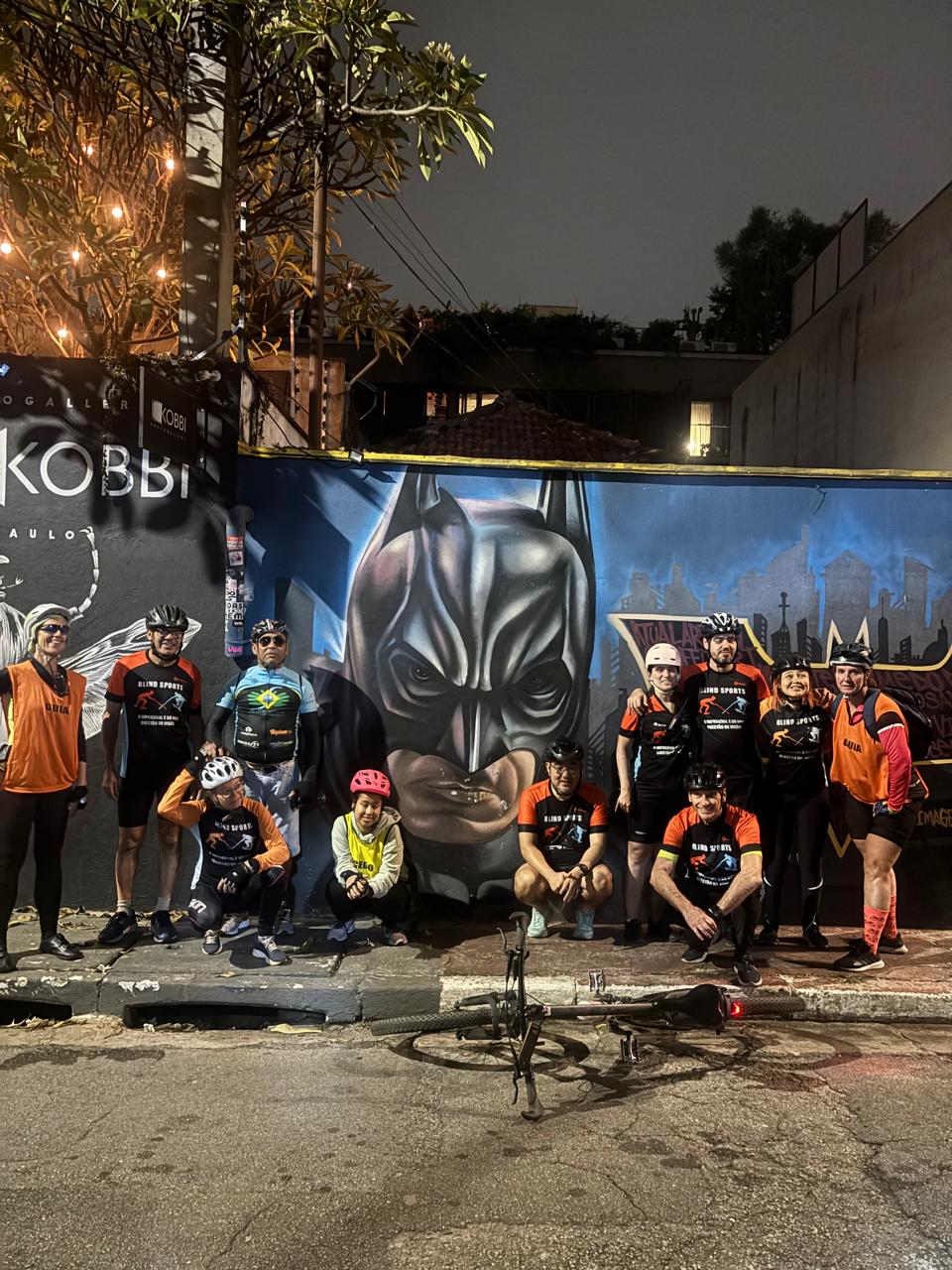 : Um grupo de pessoas com roupas de ciclismo está posando à noite na rua em frente a um mural colorido. No mural, há uma grande pintura do rosto do Batman com uma expressão séria, ao centro da imagem. À frente do grupo, há uma bicicleta preta caída no chão. As pessoas estão usando capacetes, e a maioria veste camisetas pretas com detalhes laranjas que têm a inscrição "BLIND SPORTS" e figuras de corrida e ciclismo. Dois integrantes usam coletes laranjas com a palavra "GUIA". Há árvores decoradas com luzes ao fundo, e algumas construções escuras atrás do mural.