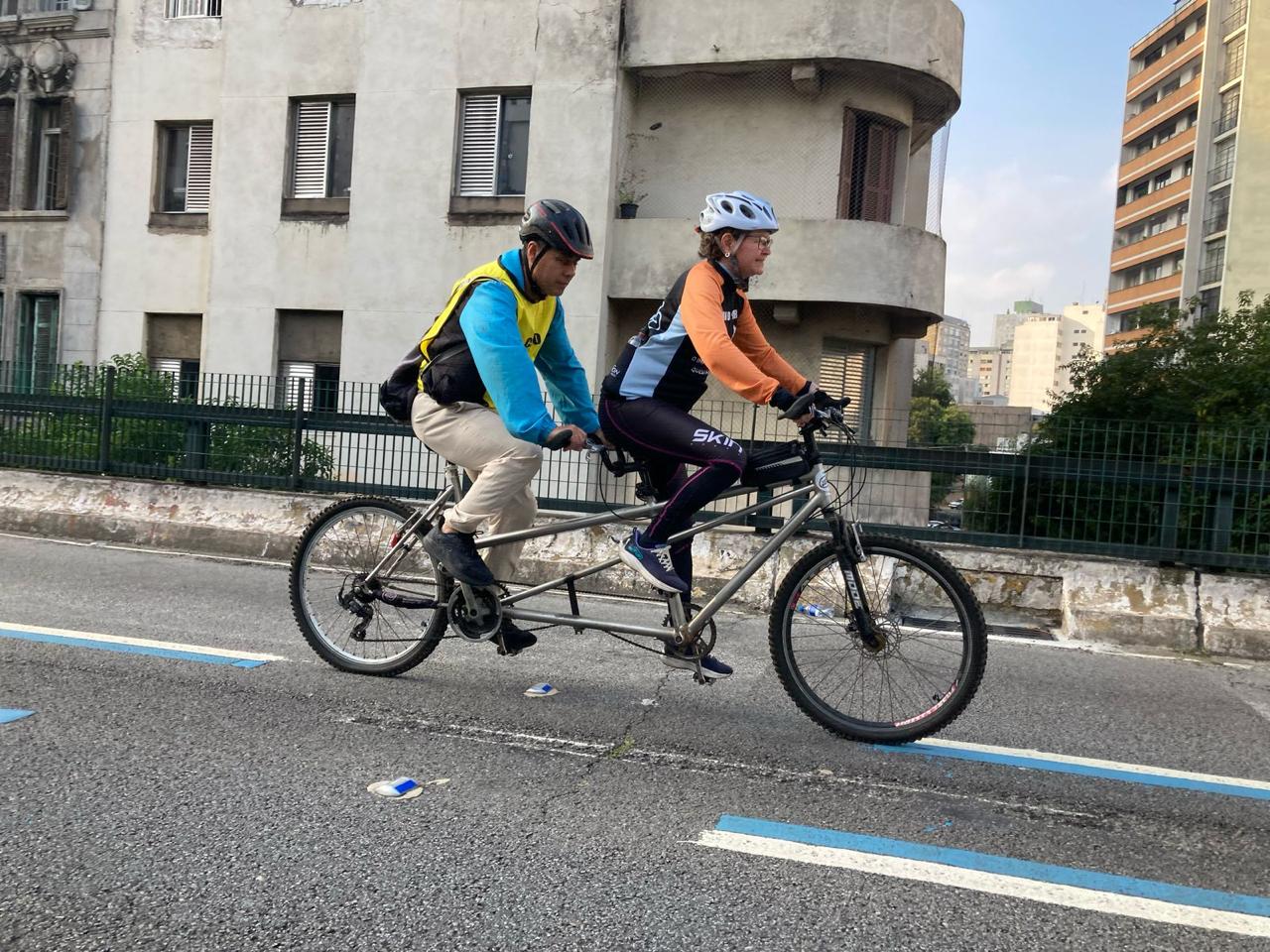 Grupo de seis pessoas posando em frente a uma fachada verde com desenho de ciclistas e o logo “ROTA 66” ao fundo. Quatro mulheres estão usando roupas esportivas de ciclismo, sendo que duas usam camisetas escritas "GUIA" e uma com "CEGO", indicando participação de guias e uma pessoa cega. Elas usam capacetes e seguram bicicletas, sendo uma delas do tipo tandem (dupla). Ao centro, um homem com roupas casuais e boné sorri para a foto. Ao lado direito, mais uma mulher ciclista usando roupas verdes e capacete. Todos parecem animados e prontos para um passeio de bicicleta inclusivo.